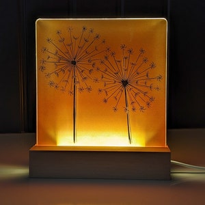 May include: A square, illuminated lamp featuring a dandelion design. The lamp has a warm, yellow glow emanating from within, with two black dandelion silhouettes. The base is made of light-colored wood, and a white cord extends from the back.