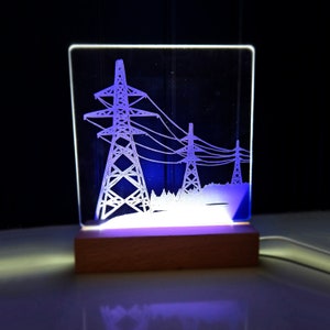 May include: A white acrylic lamp with a wooden base features an etched design of three power lines with towers. The lamp is lit with a purple light.