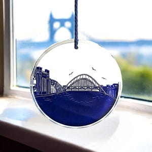 May include: A round glass bauble featuring a blue and white illustration of a bridge and cityscape. The bauble is suspended from a blue string.