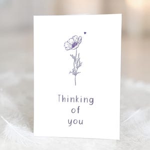 May include: A white greeting card with a purple floral illustration and the text "Thinking of you." The flower is a detailed line drawing with a heart above it. The card is simple and elegant.