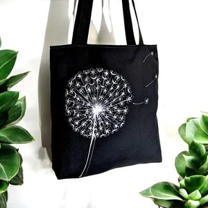May include: Black canvas tote bag featuring a white dandelion design. The dandelion has seeds blowing in the wind.