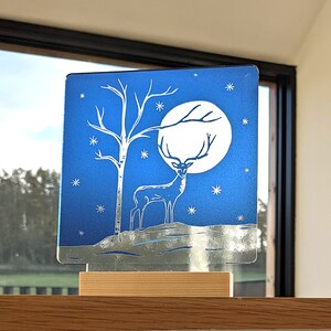 Könnte beinhalten: Eine blaue Glasscheibe mit einer silbernen Hirsch-Silhouette, die vor einem weißen Mond und einem Birkenbaum steht. Der Hintergrund ist dunkelblau mit weißen Sternen.