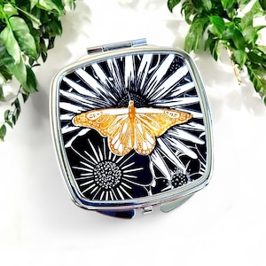 May include: A square, silver compact mirror with a black and white floral design and a gold butterfly in the center. The compact is closed and has a shiny, metallic finish. The background is white with green foliage.