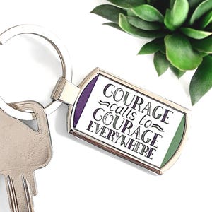Könnte beinhalten: Ein silberner Schlüsselanhänger mit einem rechteckigen Anhänger, auf dem in lila und grüner Schrift "COURAGE calls to COURAGE EVERYWHERE" auf weißem Hintergrund steht.