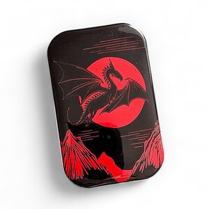 May include: Black and red tin with a dragon silhouette flying in front of a red moon. The dragon has wings spread wide and is facing the viewer. The background is black with red mountains.