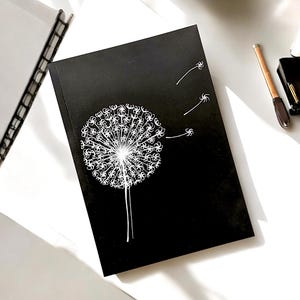Pode incluir: Um caderno preto com uma ilustração de dente-de-leão branca. O dente-de-leão tem sementes que voam ao vento.