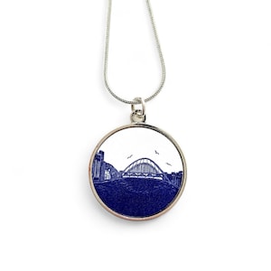 May include: A silver necklace with a round pendant featuring a blue and white illustration of a bridge over water. The pendant is surrounded by a silver border.