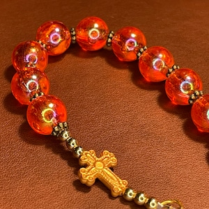 Puede incluir: Pulsera de rosario de cuentas de vidrio naranja con un dije de cruz dorada.