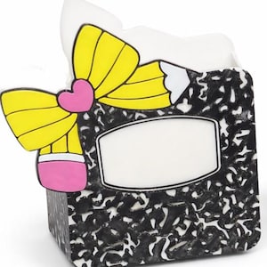 May include: A whimsical pencil-themed desk organizer. The organizer features a black and white composition notebook design with a blank label. A large yellow bow with a pink heart and pencil eraser detail adds a playful touch.