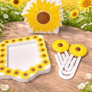 May include: A collection of white and yellow sunflower-themed home decor items. Includes a napkin holder, a picture frame, and two paper clips, all adorned with sunflower designs. The items are arranged on a wooden surface.