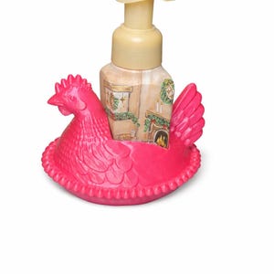 May include: A decorative soap dispenser featuring a pink chicken-shaped holder. The dispenser is beige with a pump and a painted scene of a fireplace and Christmas decorations. The chicken holder has a detailed design.