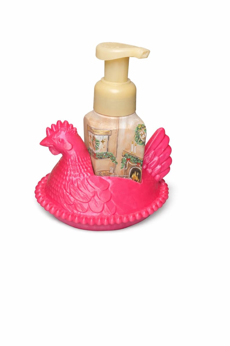 May include: A decorative soap dispenser featuring a pink chicken-shaped holder. The dispenser is beige with a pump and a painted scene of a fireplace and Christmas decorations. The chicken holder has a detailed design.