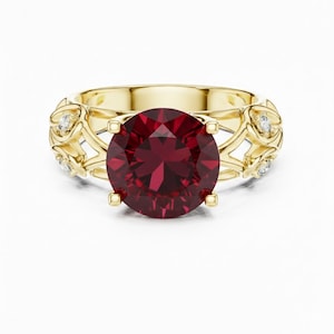 May include: A gold ring featuring a large, round, deep red gemstone. The ring has a decorative filigree design on the band, accented with small, clear gemstones. The setting is a classic four-prong style, showcasing the central stone.