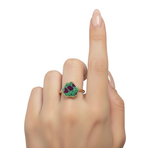 May include: A gold ring featuring a unique, irregularly shaped gemstone. The gemstone has a green base with red inclusions. The ring is displayed on a hand with a white background.