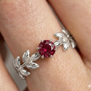 May include: A silver ring featuring a round, deep red gemstone at the center. The ring's band is designed with leaf-like details, accented with small, clear gemstones. The ring is shown on a finger.