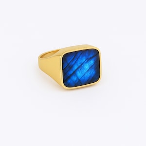 Puede incluir: Un anillo dorado con una piedra preciosa cuadrada azul. La piedra preciosa tiene un aspecto texturizado e iridiscente con tonos azules. El anillo tiene un diseño sencillo y moderno, adecuado para el uso diario.