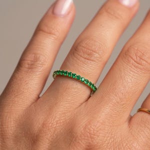 Puede incluir: Un anillo de oro con una banda de pequeñas gemas verdes redondas y vibrantes. El anillo se lleva en un dedo, con un segundo anillo de oro visible. El fondo es de un tono suave y neutro.