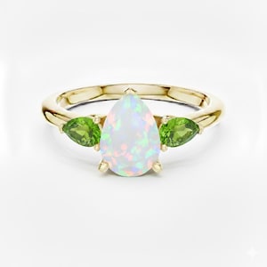 May include: A gold ring featuring a pear-shaped opal center stone, flanked by two green peridot stones. The opal has iridescent flashes of color. The ring is set on a gold band.