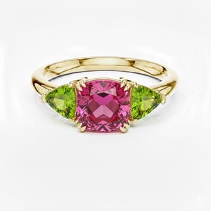 Può includere: Un anello d'oro con una grande gemma rosa quadrata affiancata da due gemme verdi triangolari. L'anello è posto su uno sfondo bianco, evidenziando i colori vivaci delle gemme.