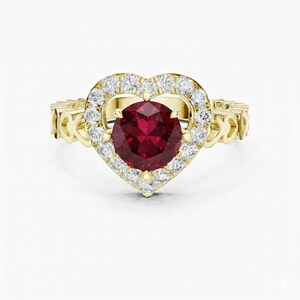May include: A gold ring with a heart-shaped design. It features a large, round, deep red gemstone at the centre, encircled by a halo of small, clear gemstones. The band is also decorated with ornate details.