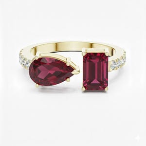 May include: A gold ring featuring a pear-shaped and emerald-cut red gemstone design. The ring band is adorned with small, clear gemstones. The ring is set against a white background.
