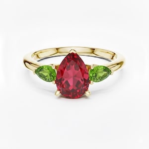May include: A gold ring featuring a large, pear-shaped, red gemstone flanked by two smaller, green, pear-shaped gemstones. The ring has a simple, elegant design, perfect for a special occasion.