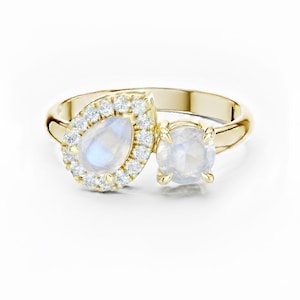 Könnte beinhalten: Ein goldener Ring mit zwei Mondstein-Edelsteinen. Einer ist birnenförmig und von kleinen, klaren Steinen umgeben. Der andere ist rund. Der Ring hat ein poliertes Goldband und ist vor weißem Hintergrund platziert.