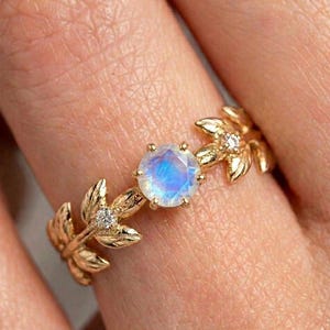 Peut inclure: Bague dorée ornée d'une pierre précieuse ronde et irisée au centre. L'anneau est orné de motifs en forme de feuilles et de petits accents scintillants, créant une esthétique élégante et inspirée de la nature.