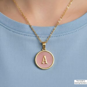 May include: Gold-tone necklace with a pendant displaying the letter "A" in gold against a pink backdrop. The circular pendant has a gold border. The necklace is shown on a light blue top.