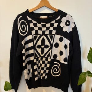 May include: A long-sleeve black and white knit sweater featuring a crew neckline and a patchwork-style design. The pattern includes alternating sections of checkerboard, polka dots, stripes, floral motifs, and spiral shapes in a high-contrast knit.