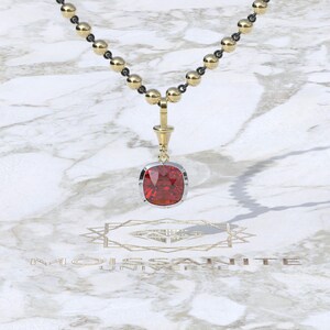 May include: A gold-toned necklace with a pendant featuring a large, square-cut, red gemstone. The gemstone is set in a silver-colored bezel. The necklace chain has alternating gold and black beads. The background is a marble-like surface.
