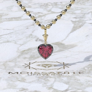 Puede incluir: Un collar dorado con un colgante en forma de corazón. El colgante presenta una piedra preciosa roja engastada en un marco de metal oscuro. La cadena del collar está compuesta por alternancia de cuentas doradas y eslabones negros. La marca "MOISSANITE UNIVERSE" es visible.