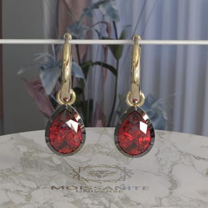 Pode incluir: Um par de brincos de argola dourados com pedras preciosas vermelhas facetadas em forma de gota. As pedras são fixadas em uma moldura de metal escuro. Os brincos são exibidos em uma barra branca.
