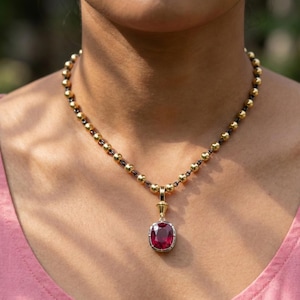 Puede incluir: Un collar de cuentas doradas con un colgante con una gran gema roja rectangular. El colgante está enmarcado en un engaste plateado con detalles dorados. El collar se lleva sobre una blusa rosa.