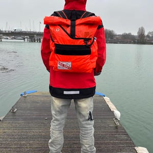 May include: An orange and black backpack with the text "LIFE RAFT" is worn over a red jacket. The person is wearing gray jeans and black and white sneakers. The background shows a body of water and a bridge.