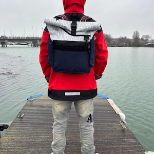 Puede incluir: Una persona con una chaqueta roja con capucha y vaqueros grises claros está de pie en un muelle de madera, de espaldas a la cámara. Se lleva una mochila con un estampado blanco y gris y una parte inferior azul marino. Zapatillas deportivas blancas y negras completan el atuendo.