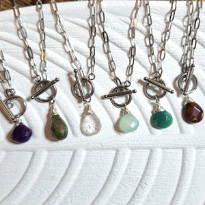 May include: Seven silver necklaces with toggle clasps and various gemstone pendants. The gemstones include amethyst, green, clear, light green, and red.