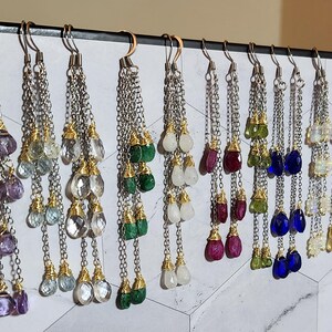 May include: A collection of dangling earrings with various gemstone colors, including purple, green, clear, and blue. Each earring features a chain design with multiple teardrop-shaped stones and gold-colored accents. The earrings are displayed on a white and gray patterned surface.