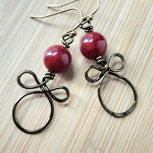 May include: A pair of dangle earrings featuring round, red, marbled beads suspended from antique brass-toned wire. Each earring showcases a decorative wire-wrapped loop design at the base, finished with a simple hook for ear attachment.