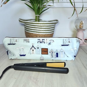 May include: A light blue zippered pencil case with a beach hut and sailboat pattern. The case has a brown leather tag that reads "J Shack". A black and yellow flat iron is in front of the case.