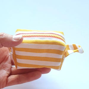 May include: A small, yellow and white striped zippered pouch held in a hand. The pouch has a matching fabric loop on one end and a coral-coloured zip. The fabric is a soft, lightweight material.