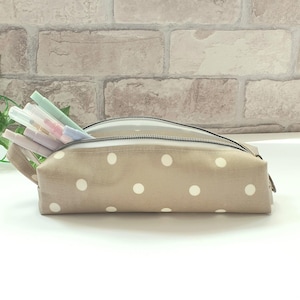May include: A beige pencil case with white polka dots, partially unzipped, showing colourful pens. The case has a zip closure and a small fabric loop. The background is a light-coloured brick wall.