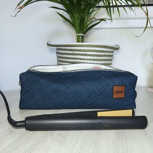 May include: A black hair straightener with gold-colored plates rests on a white surface. A dark blue denim-textured pencil case with a brown leather tag is behind it. A green and white striped basket and a potted plant are in the background.