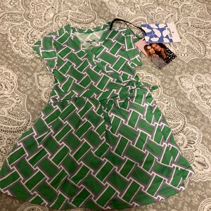 May include: A green and purple geometric patterned wrap dress with short sleeves. The dress has a V-neckline and a tie at the waist. The dress is made by the brand "The Little Twig".