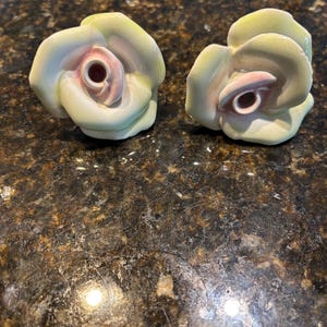 May include: Two ceramic rose-shaped cabinet knobs. The knobs feature a pale yellow and white color scheme with pink accents in the center. The knobs are designed to resemble open roses.