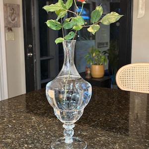 May include: A clear glass vase with a red rose and green leaves. The vase has a bulbous body and a round base. The vase is sitting on a dark brown speckled surface. The background includes a door and a chair.