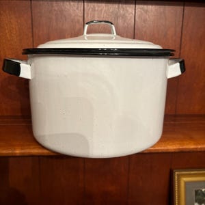 May include: White speckled enamelware pot with a black rim and handles. The pot has a matching lid with a black handle. The pot is sitting on a wooden shelf.