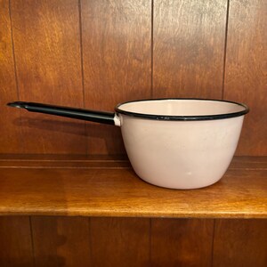May include: A pink enamel saucepan with a black handle. The saucepan has a black rim around the top.