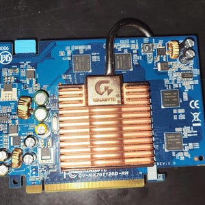 Peut inclure: Une carte graphique bleue Gigabyte GV-NX76T128D-RH avec un dissipateur thermique en cuivre et un caloduc argenté. La carte comporte divers composants électroniques et connecteurs.