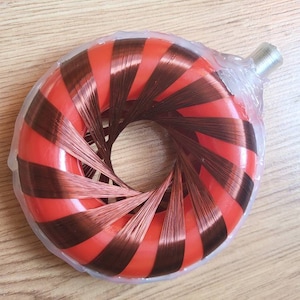 Puede incluir: Un inductor toroidal rojo y marrón, envuelto en plástico transparente. El inductor tiene forma circular con rayas rojas y marrones alternas. Un conector plateado sobresale del lateral.